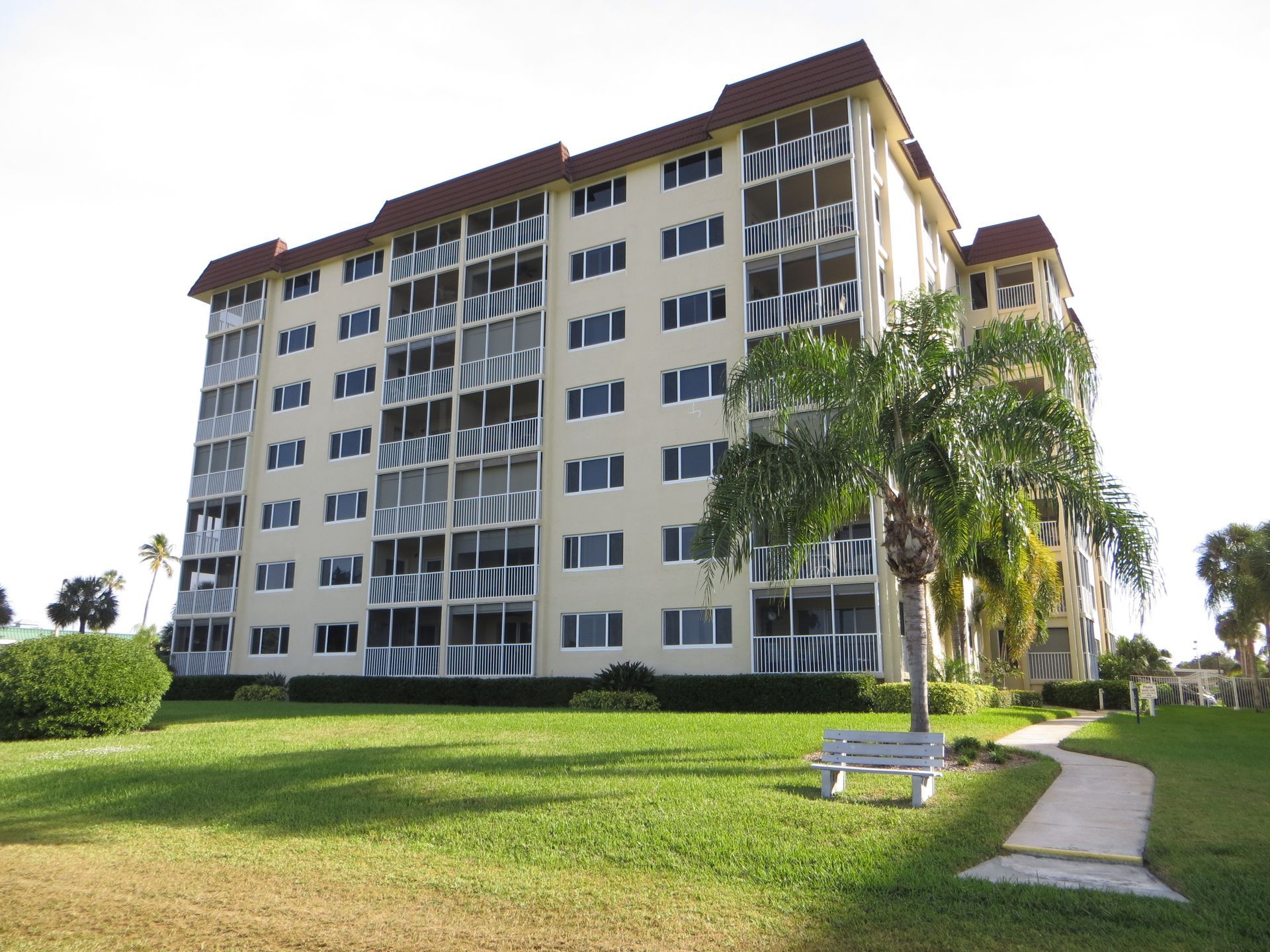 A multi-story beige apartment building with screened balconies, surrounded by a lawn, a palm tree, and a sidewalk.