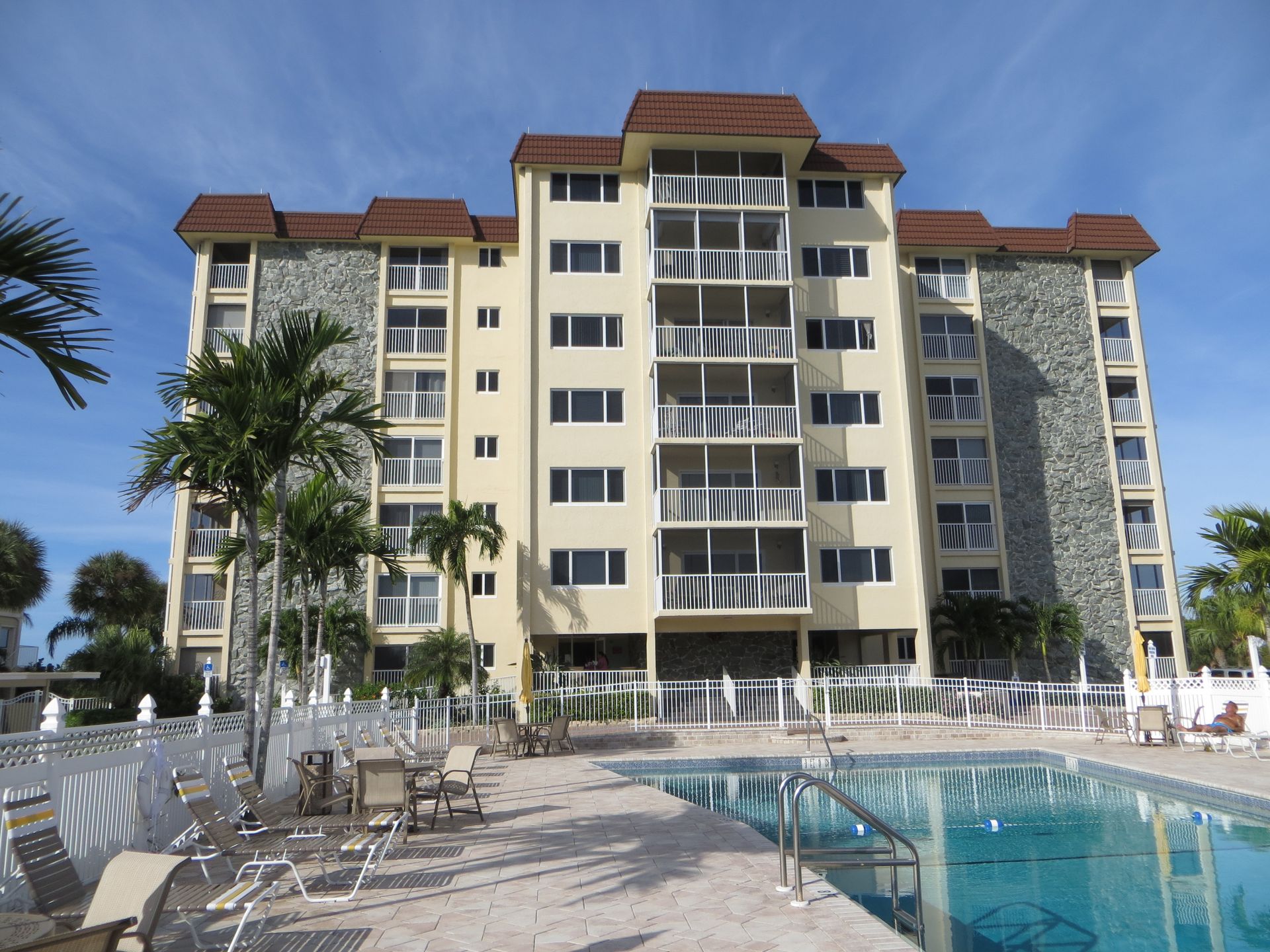 A multi-story condo building with balconies and stone siding, overlooking a swimming pool and patio area.