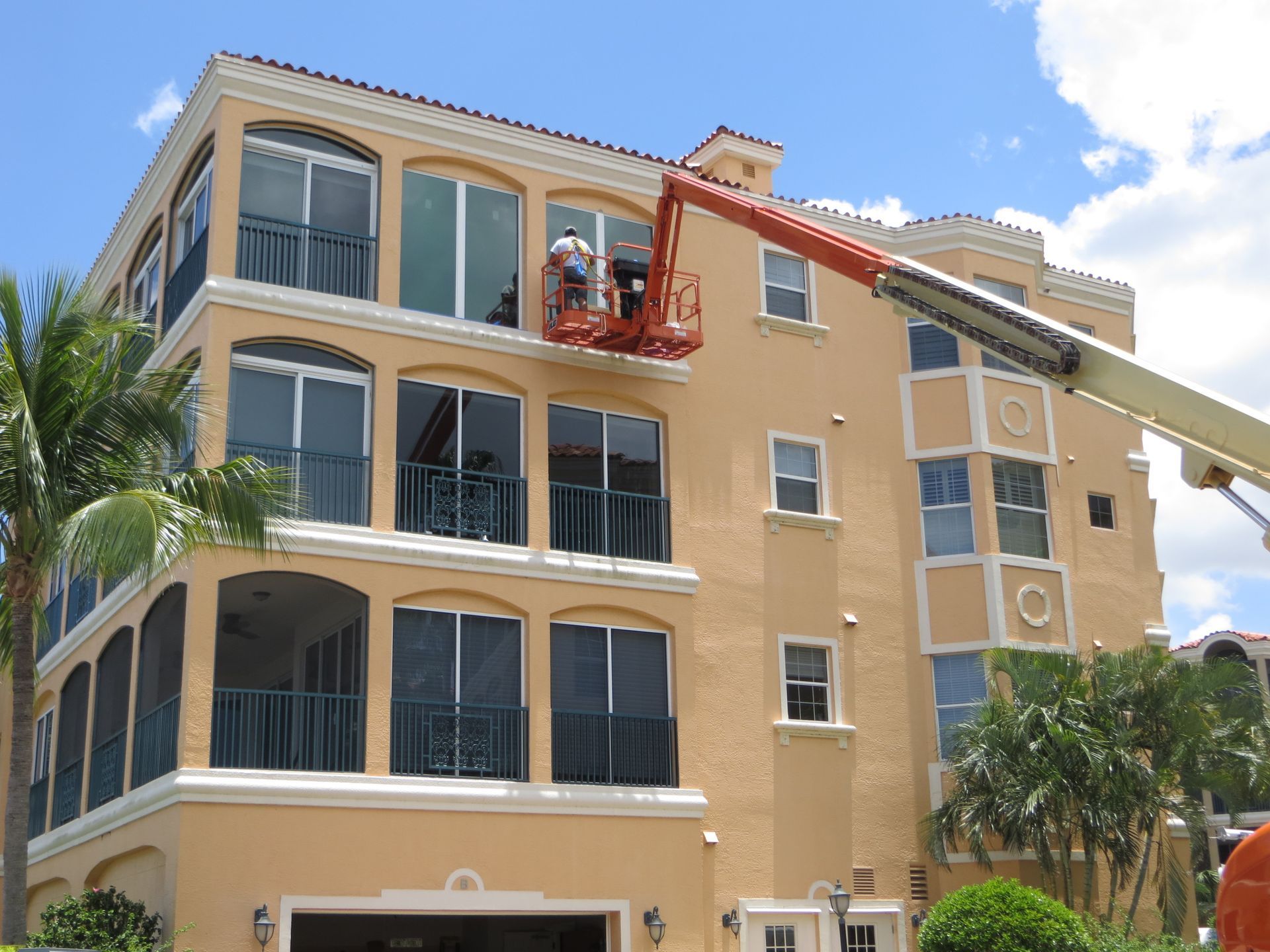 Two workers in a lift platform clean the upper-story windows of a beige, multi-story building under a blue sky.
