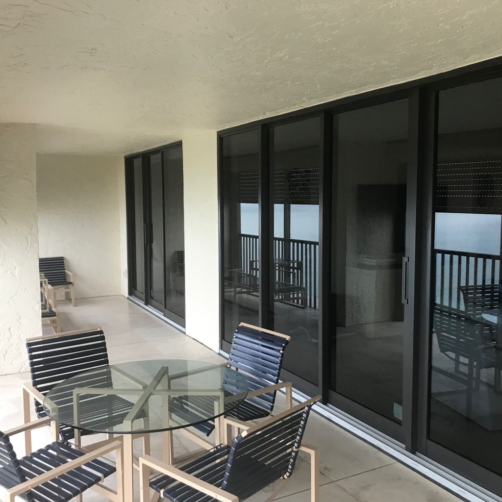 A glass-topped dining table with four dark chairs sits on a covered balcony next to a row of sliding glass doors.