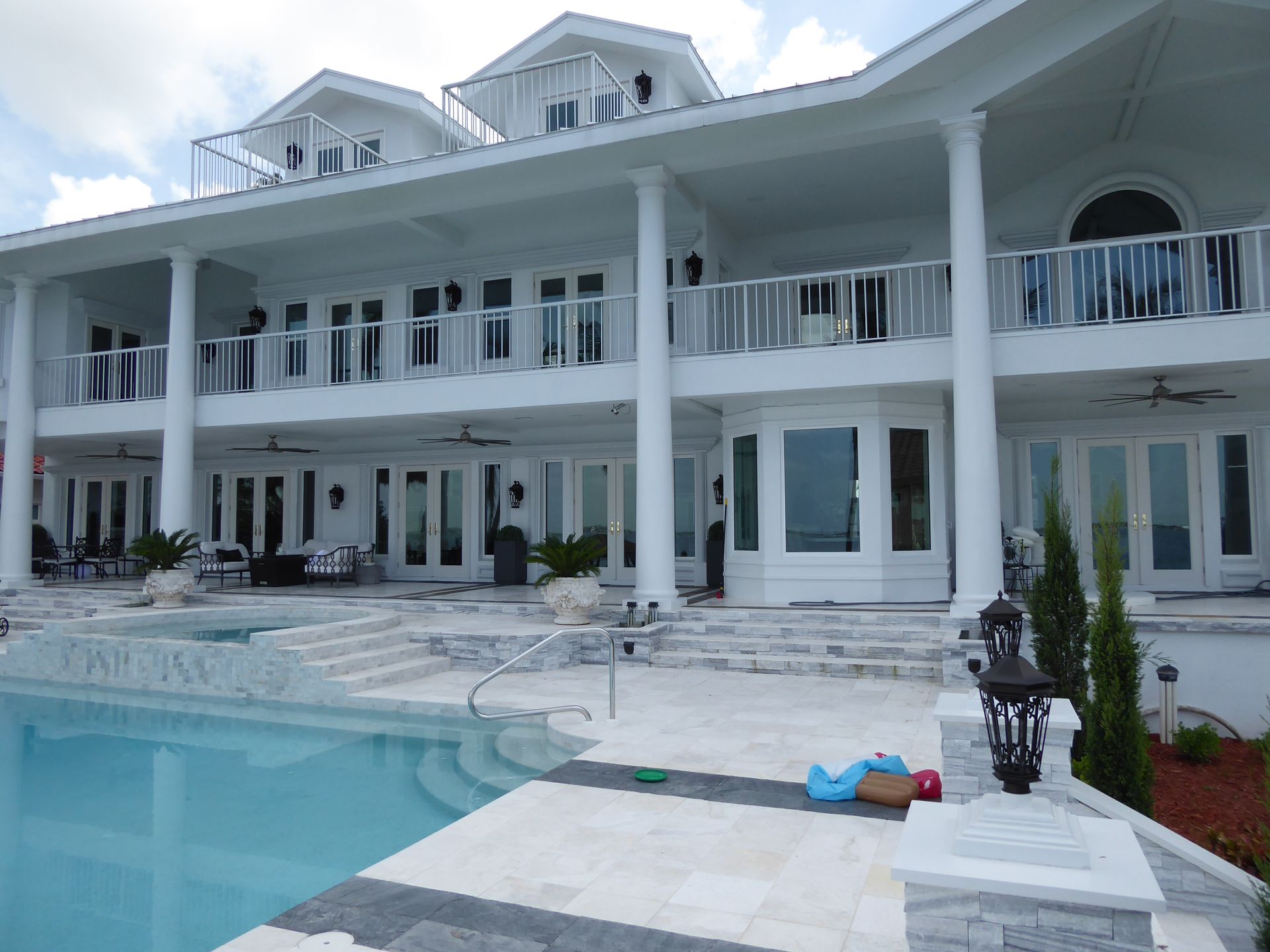 A large, two-story white house with tall columns, balconies, and a patio surrounding an outdoor swimming pool.