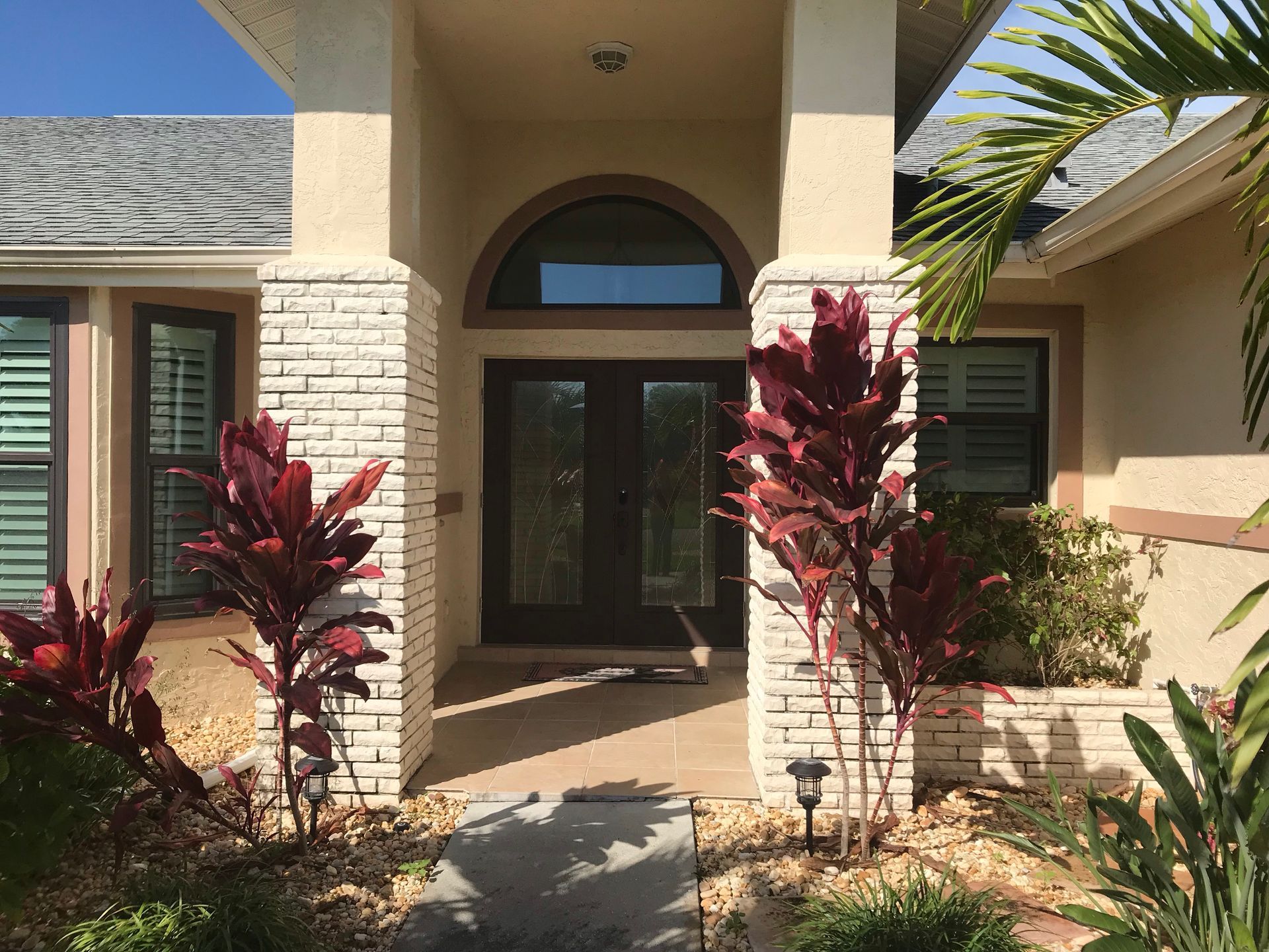 Front entrance of a house with a dark double door, arched window, and two brick pillars flanked by red-leafed plants.
