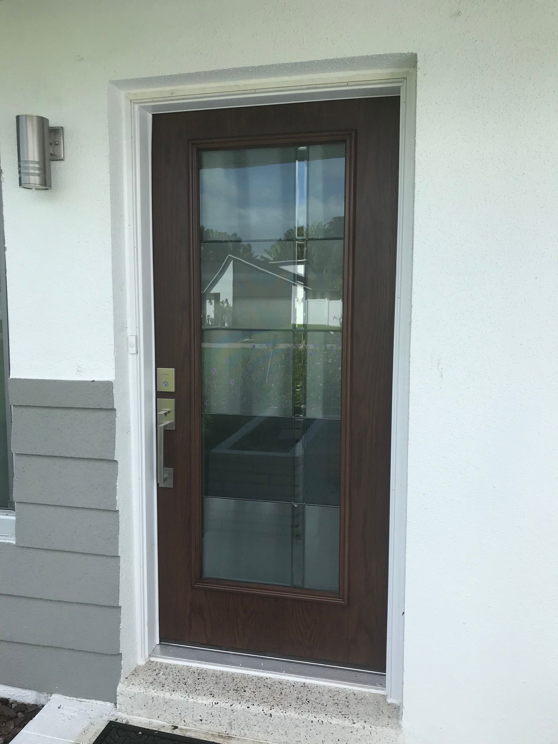 A dark brown exterior door with a long glass window panel, set in a white wall with gray siding nearby.