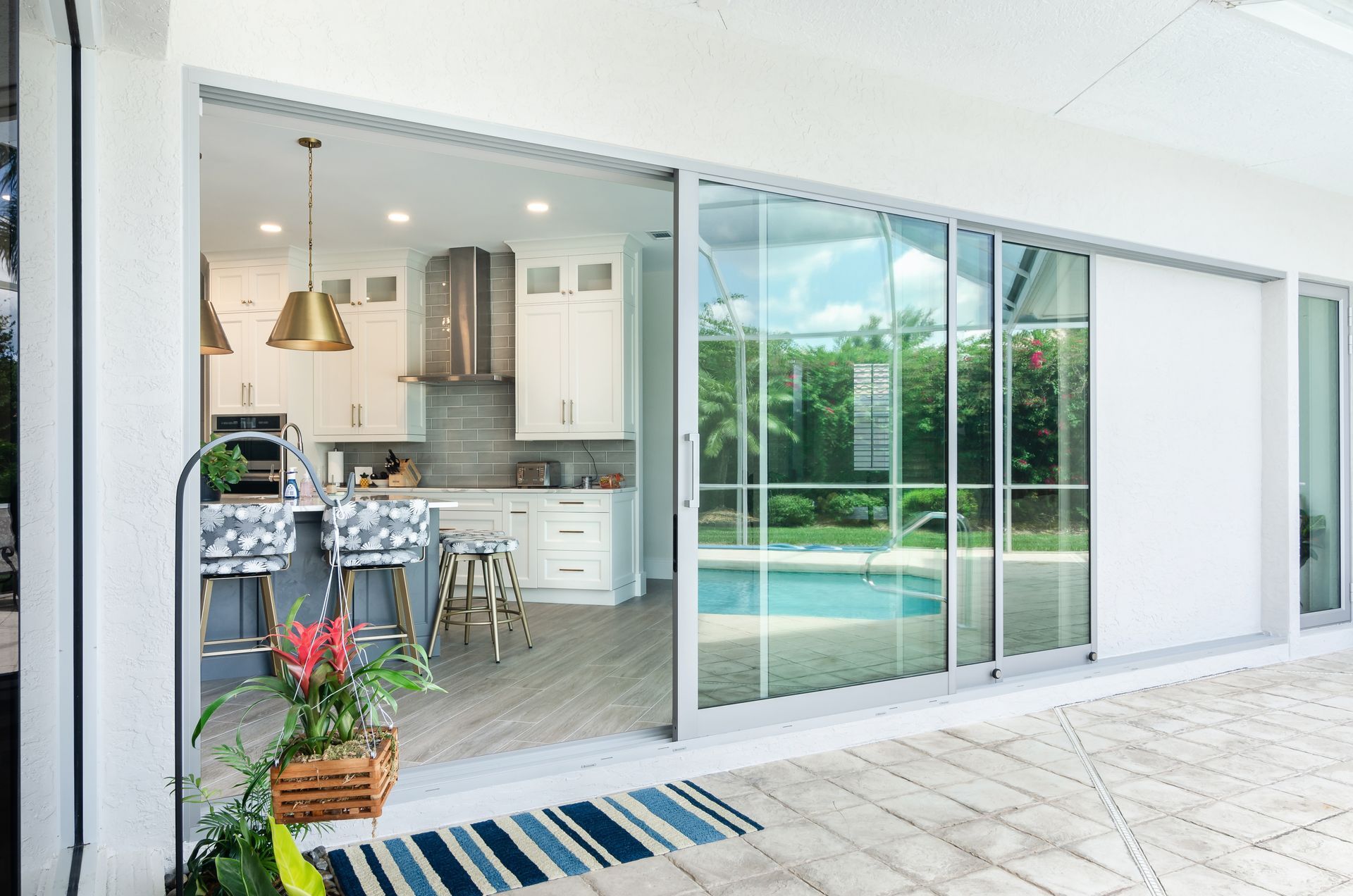 A modern kitchen viewed through wide glass sliding doors, showing an island, stools, and a pool area on the patio.