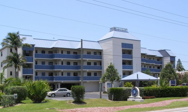A multi-story beige and blue residential apartment complex with a palm tree, landscaping, and a car in the parking lot.