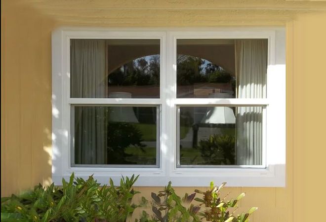 A white-framed window on a pale yellow exterior wall, showing a partial reflection of a house and trees.