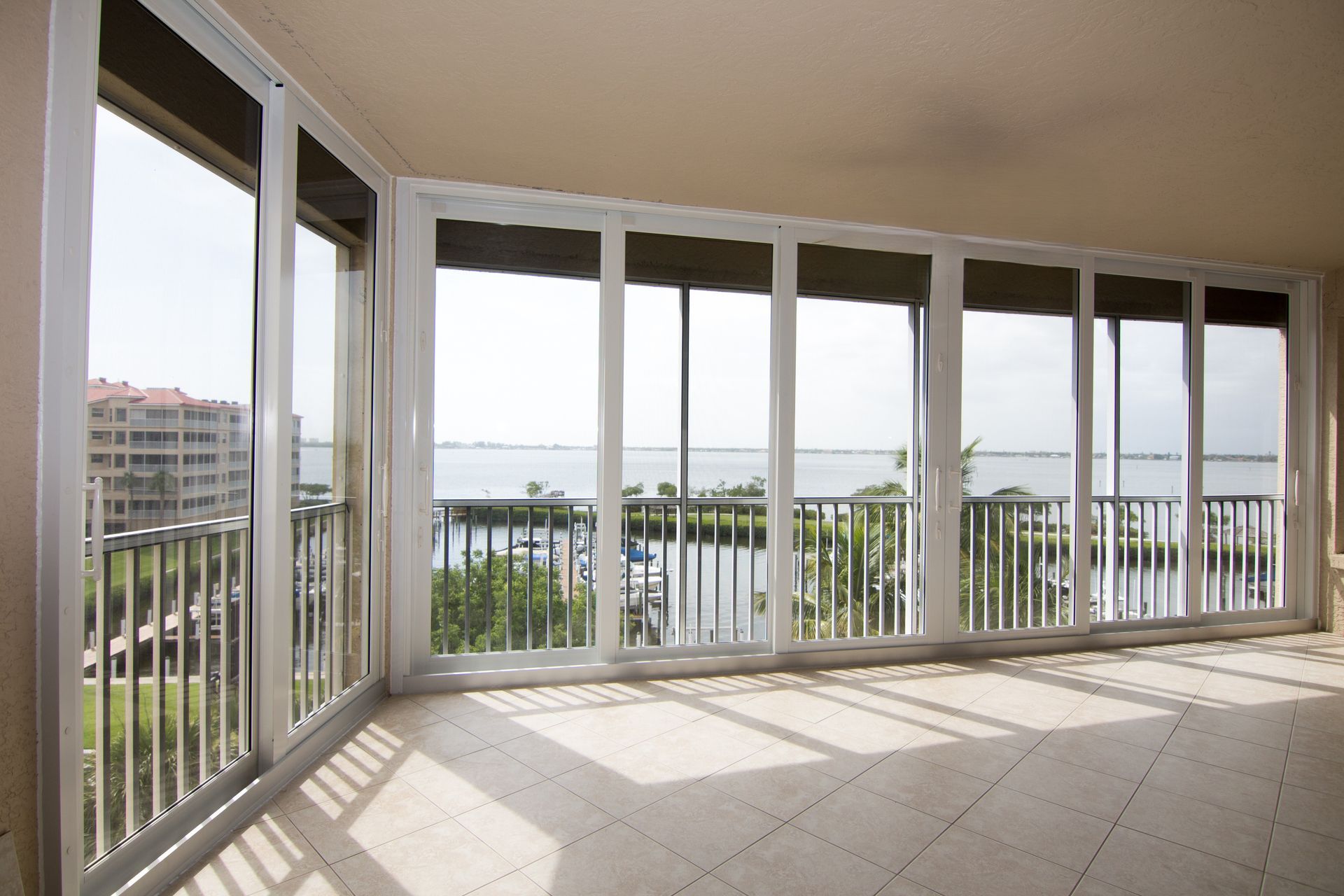An indoor balcony with large glass windows overlooking a body of water, a marina, and buildings on a sunny day.