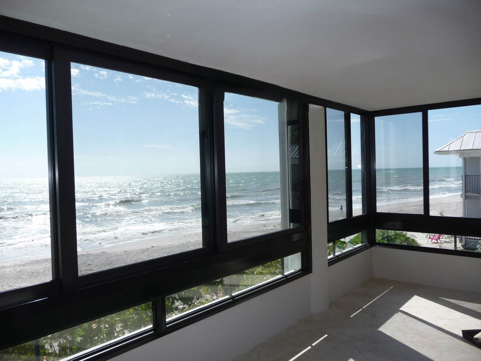 Bright ocean view through large windows from a sunlit room with dark frames and a balcony.