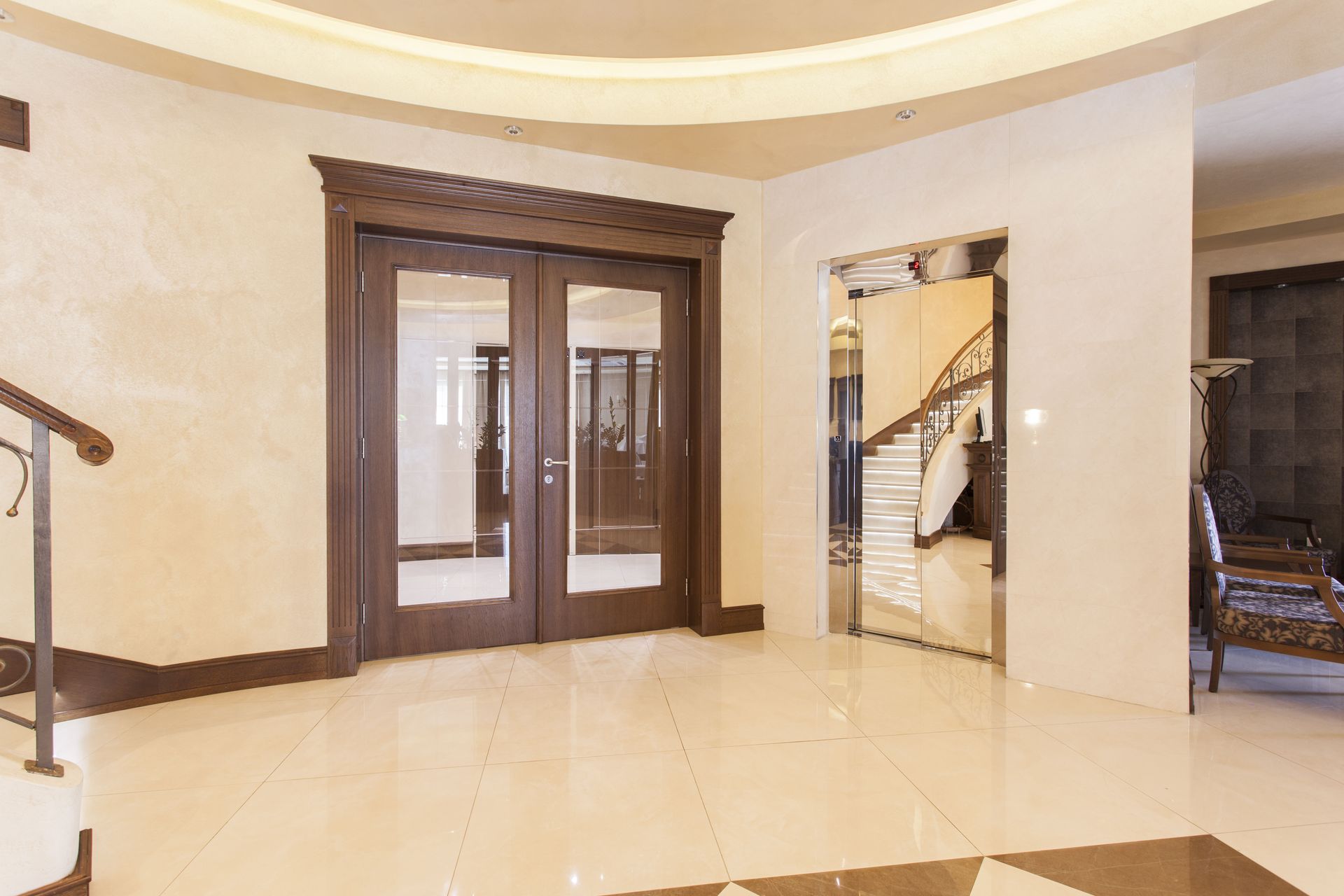 A bright foyer features a dark wood double door, a glass-encased staircase reflecting in a mirror, and tiled flooring.