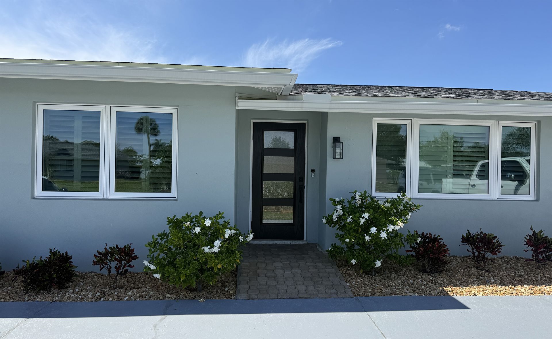 Single-story gray house with white-trimmed windows, a black front door, and two flowering shrubs in front.