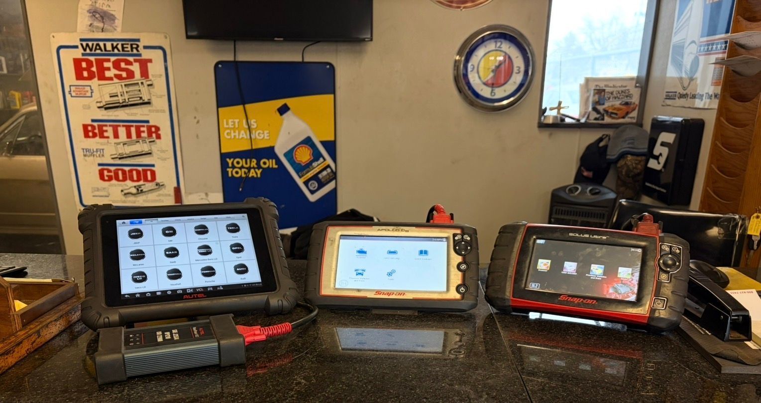 Three diagnostic devices on a workbench in an auto shop. Display screens show vehicle system icons. Shop decorations in background.