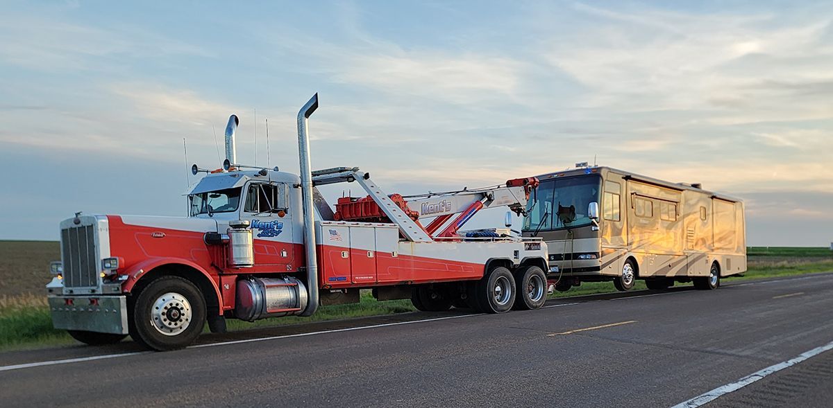 Red and white tow truck hauling a damaged car on a flatbed beside a yellow trailer on a roadside