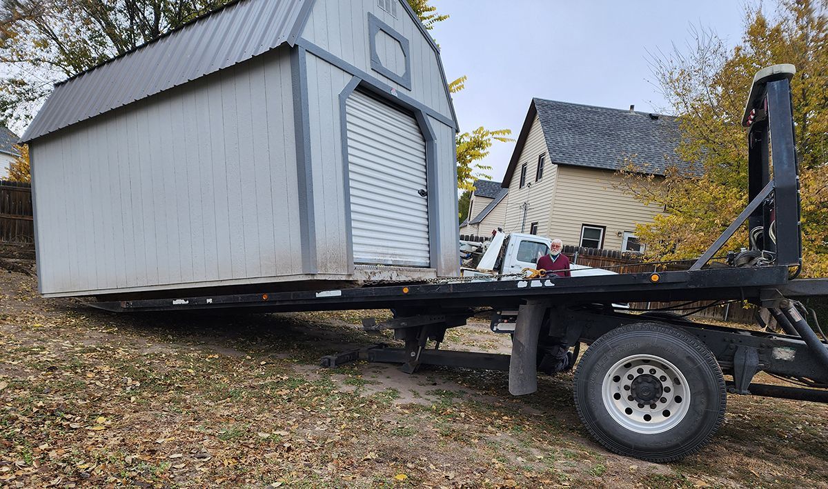 Small gray shed loaded on a utility trailer in a yard, tilted with a ramp at the back