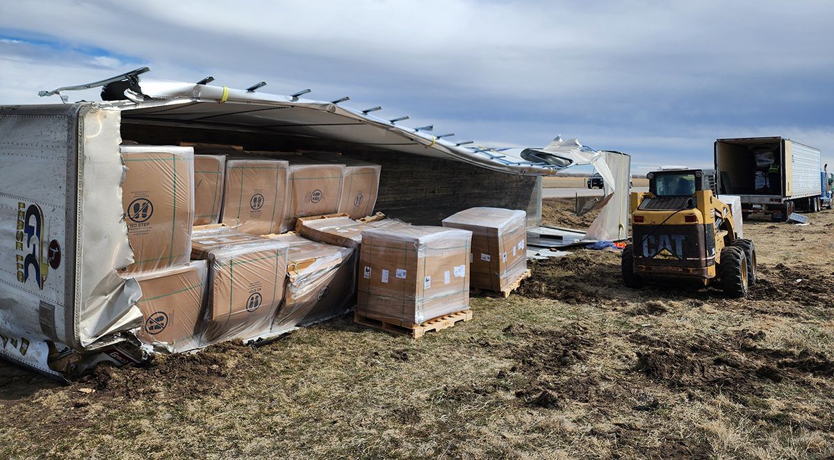 Overturned shipping container with damaged cargo boxes and a forklift in a muddy field