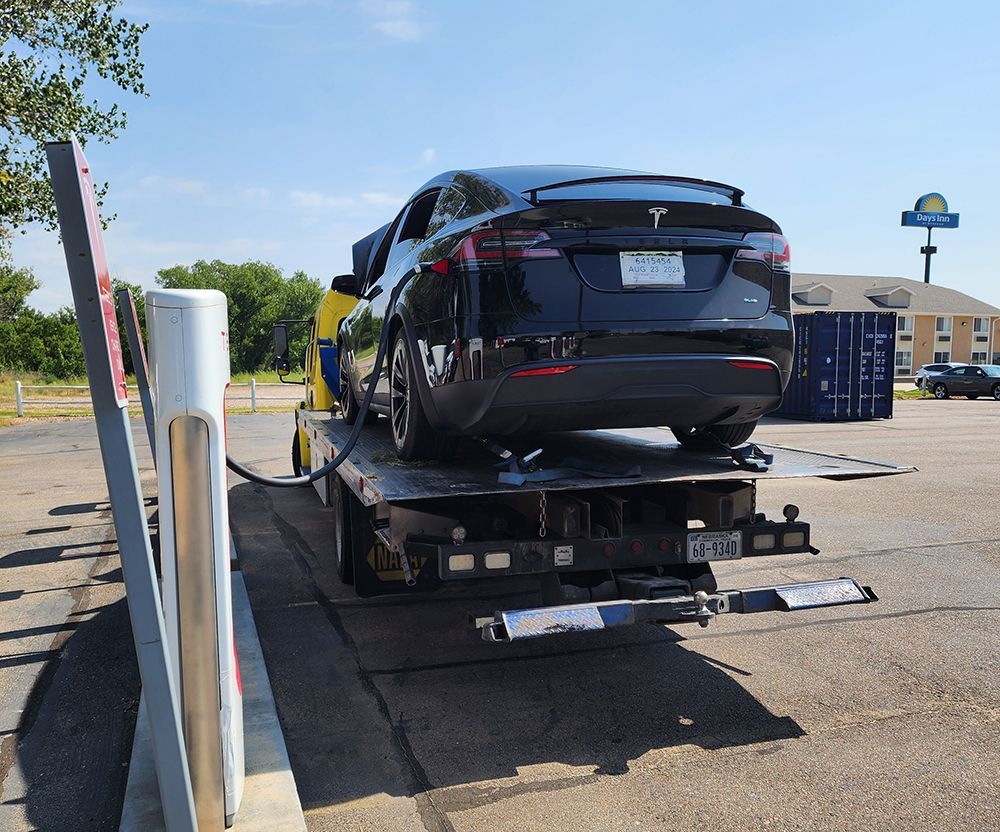 Black Tesla on a flatbed tow truck in a parking lot beside a charging station