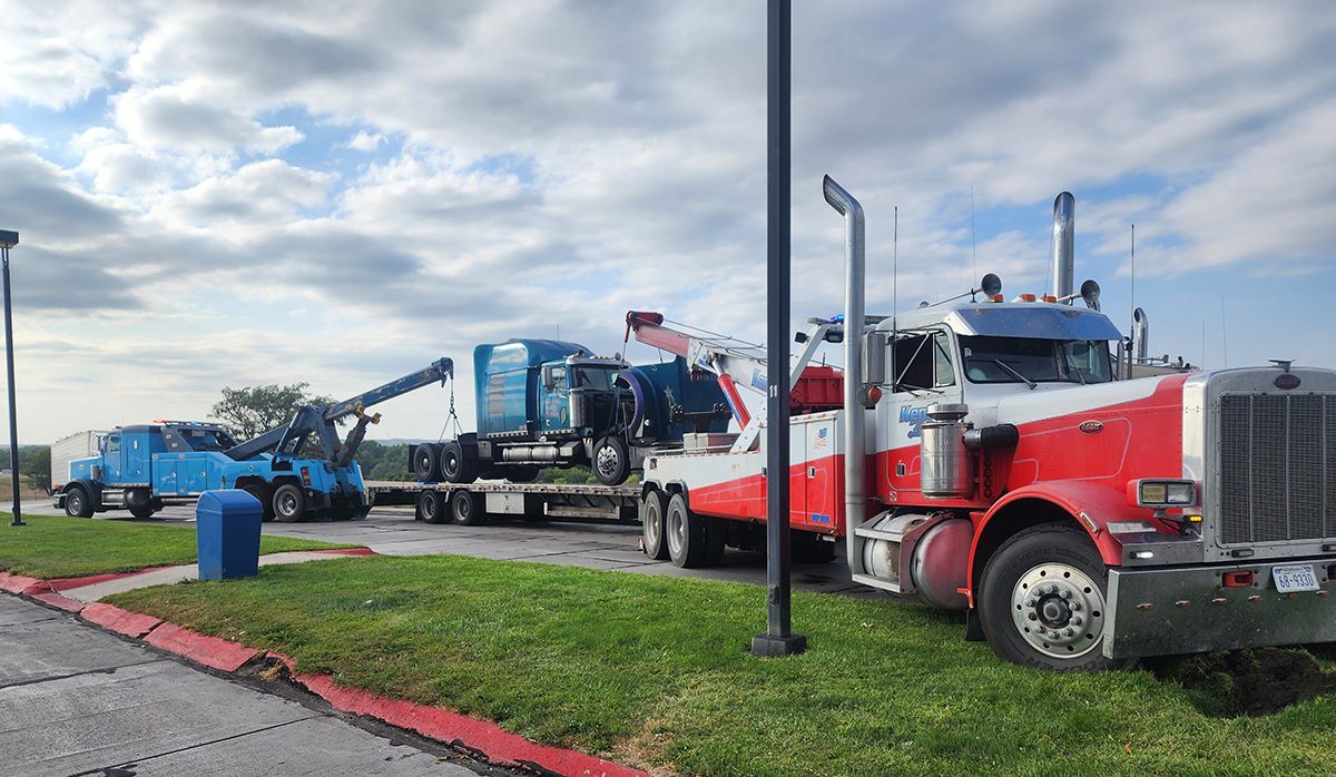 Tow trucks lined up roadside, with a red truck in front and a blue truck behind on a cloudy day