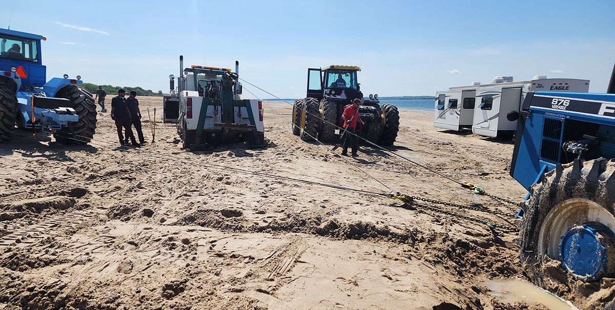Tractors and beachgoers on a sunny sandy beach, with parked vehicles along the shoreline.