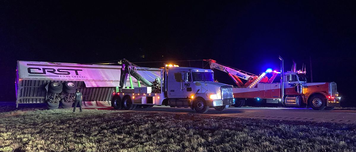 Tow truck and emergency vehicles beside a damaged semitrailer at night in a field