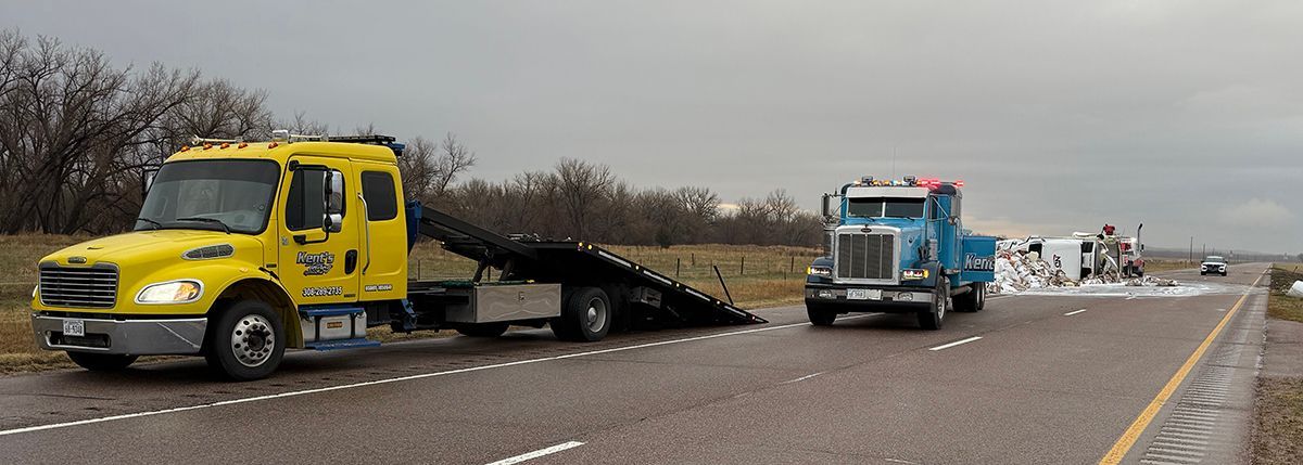 Yellow tow truck hauling a car on a highway beside a blue truck under cloudy skies