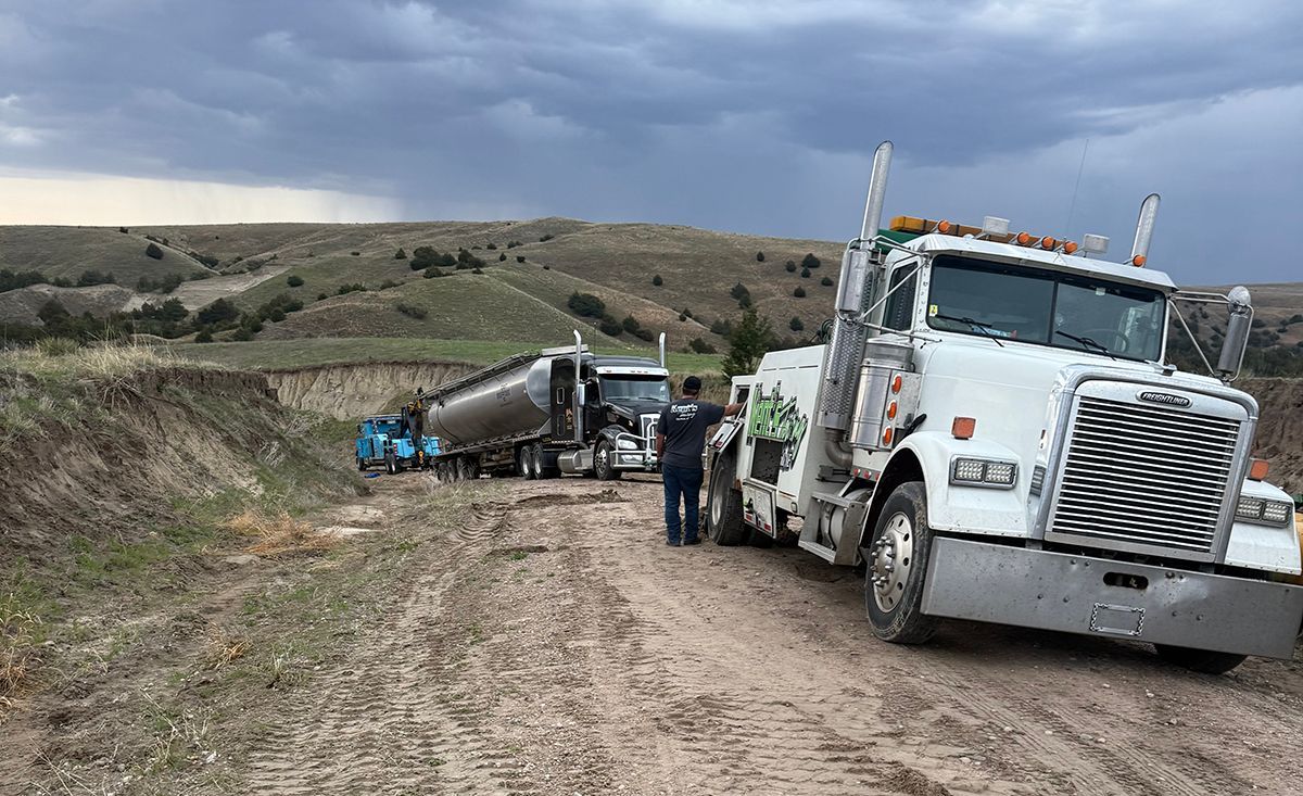 Tow truck and semi-trailer on a dirt road in a rural field under cloudy skies