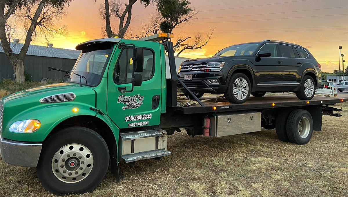 Green tow truck carrying a black SUV on a flatbed at sunset