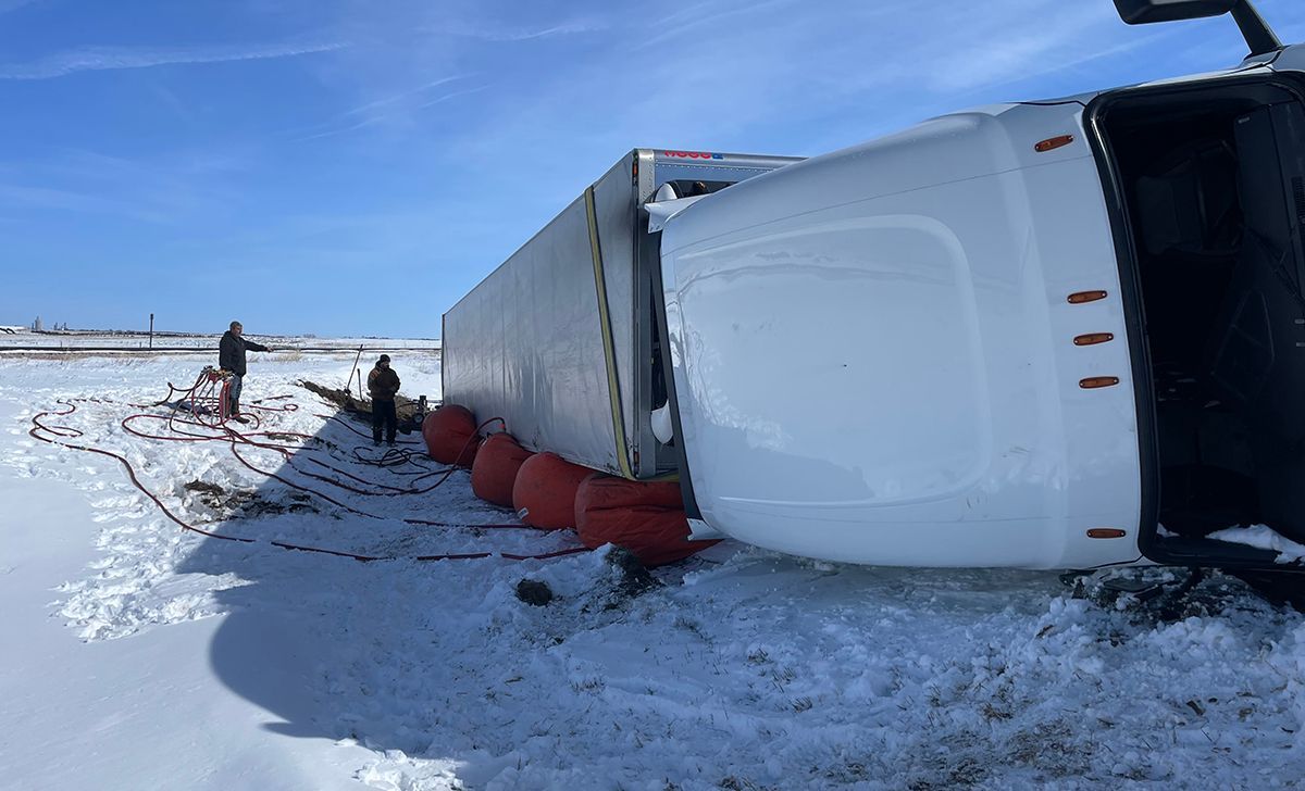 Overturned semi-truck on snowy roadside with red barriers and a person walking nearby