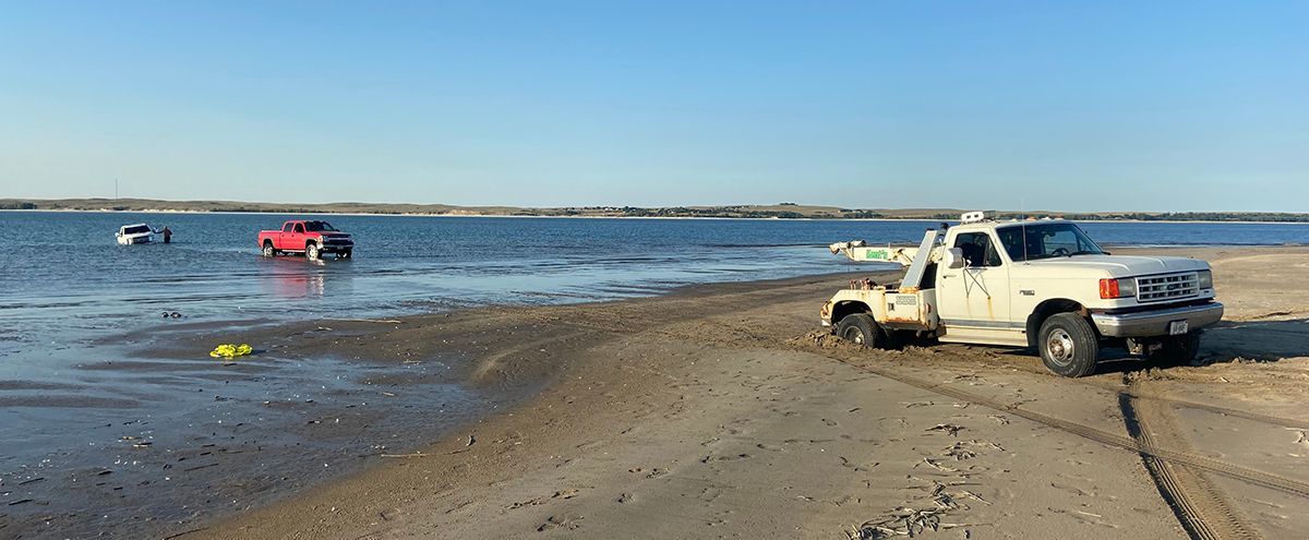 White pickup truck parked on a sandy beach near calm water, with a red boat in the distance.