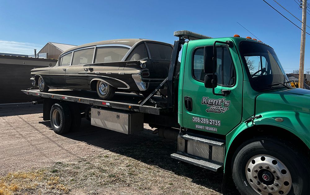Green tow truck carrying a black vintage sedan on a flatbed under a clear sky