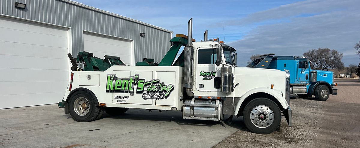 White tow truck with green logo parked outside a warehouse, with a blue truck in the background.