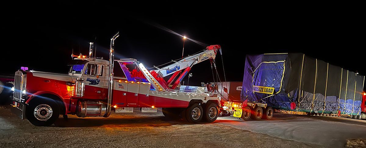 Red tow truck loading a crashed blue semi-trailer at night on a gravel road