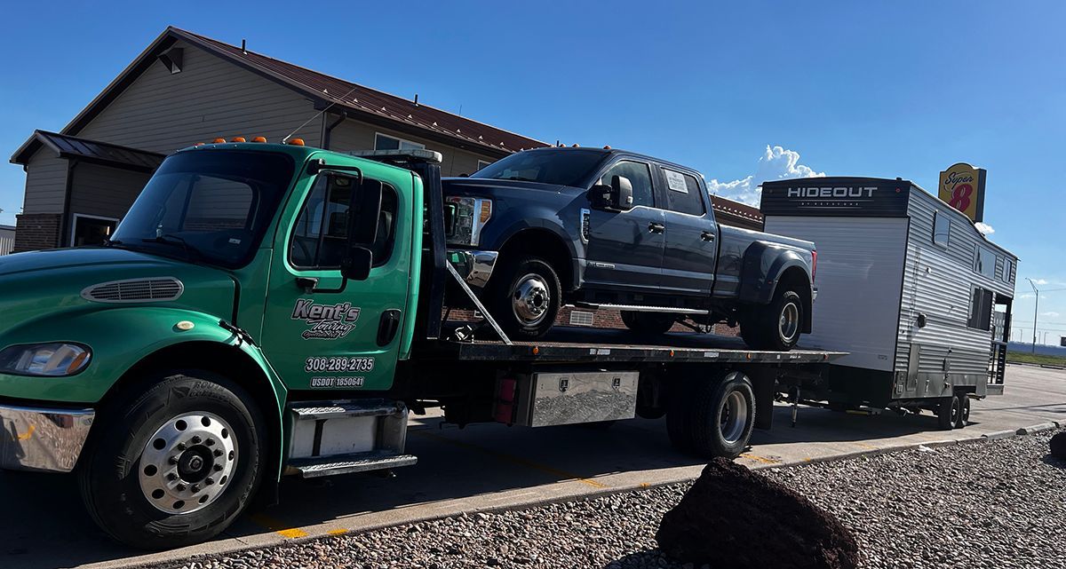 Green tow truck carrying a black SUV and a silver trailer on a gravel lot near a building