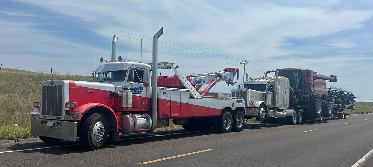 Red tow truck hauling a large black heavy-duty vehicle on a roadside under a clear sky