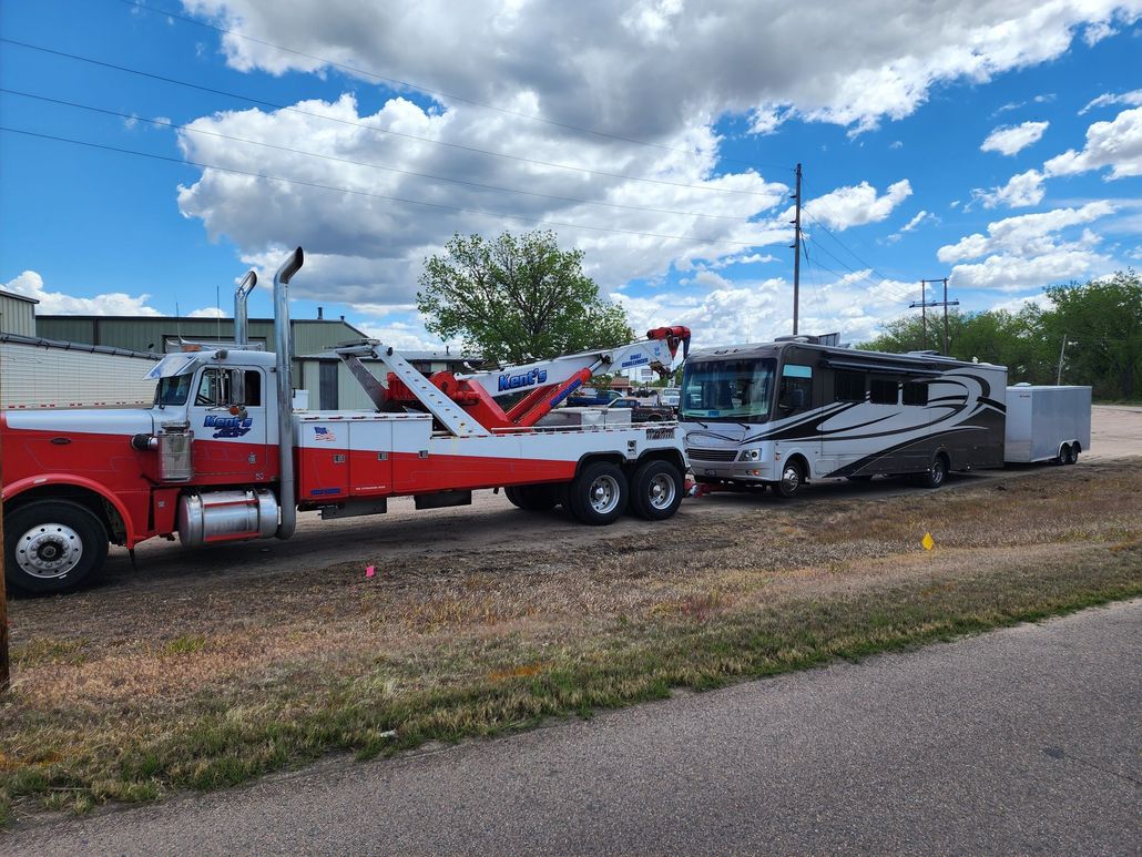 Red tow truck hauling a damaged silver car on a trailer beside a roadside under a blue sky
