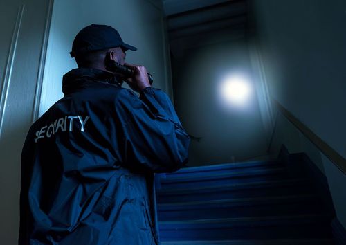 Security guard ascending dim stairs with flashlight, scanning a dark hallway