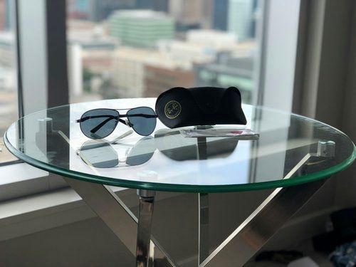 A pair of sunglasses and a case are on a glass table in front of a window.