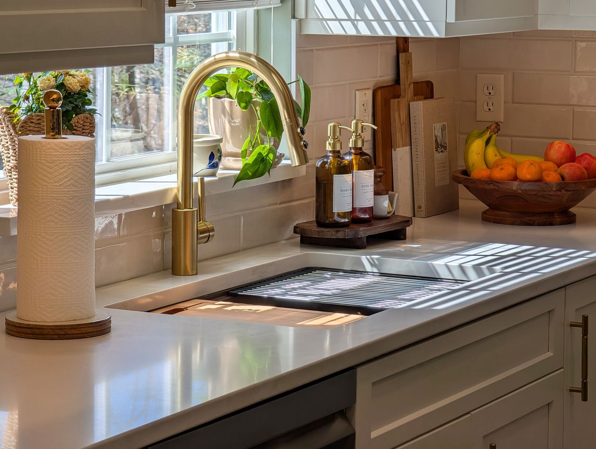 Kitchen countertop with gold faucet, sink, and dish rack. Window with plants, fruit bowl, and paper towel holder.