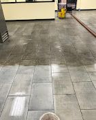 Wet, gray tiled floor being cleaned in a commercial space, with cleaning supplies visible.
