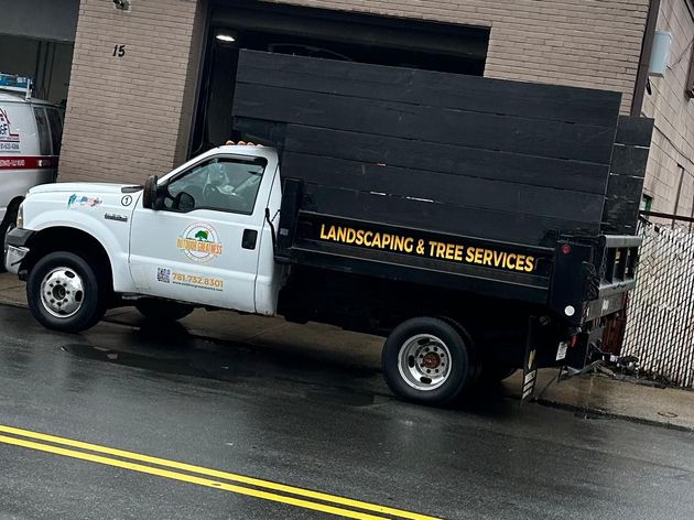 White landscaping truck with black dump bed parked on a wet street beside a building.