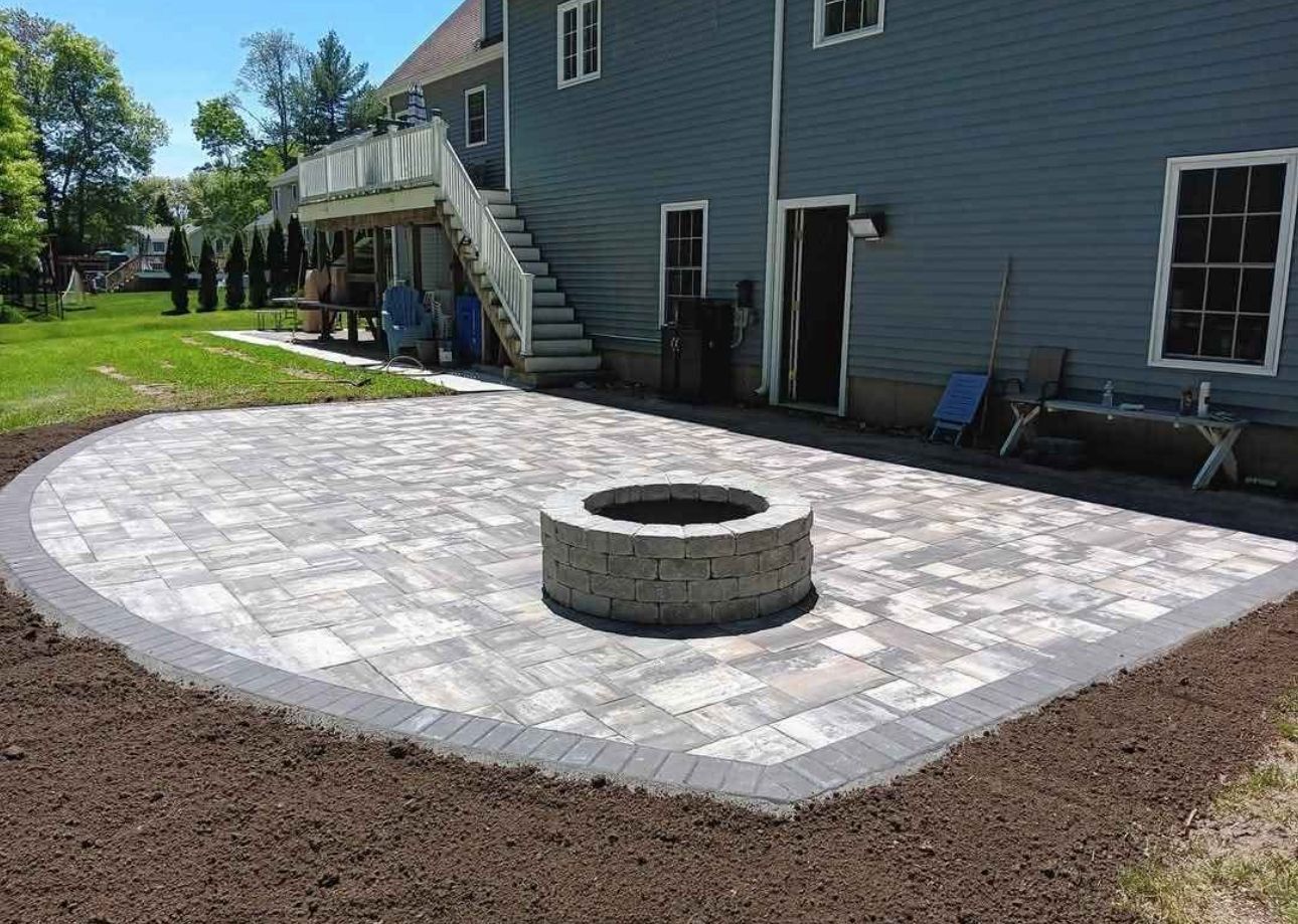 Backyard patio with circular fire pit and light stone pavers beside a blue house