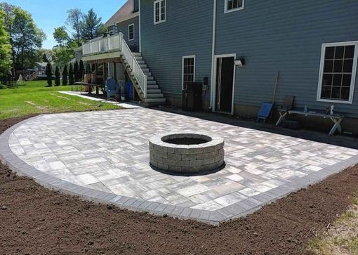 Backyard patio with circular fire pit and light stone pavers beside a blue house