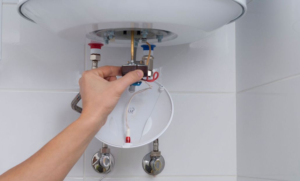 Hand adjusting the electrical connections on a water heater, indoors against a tiled wall.