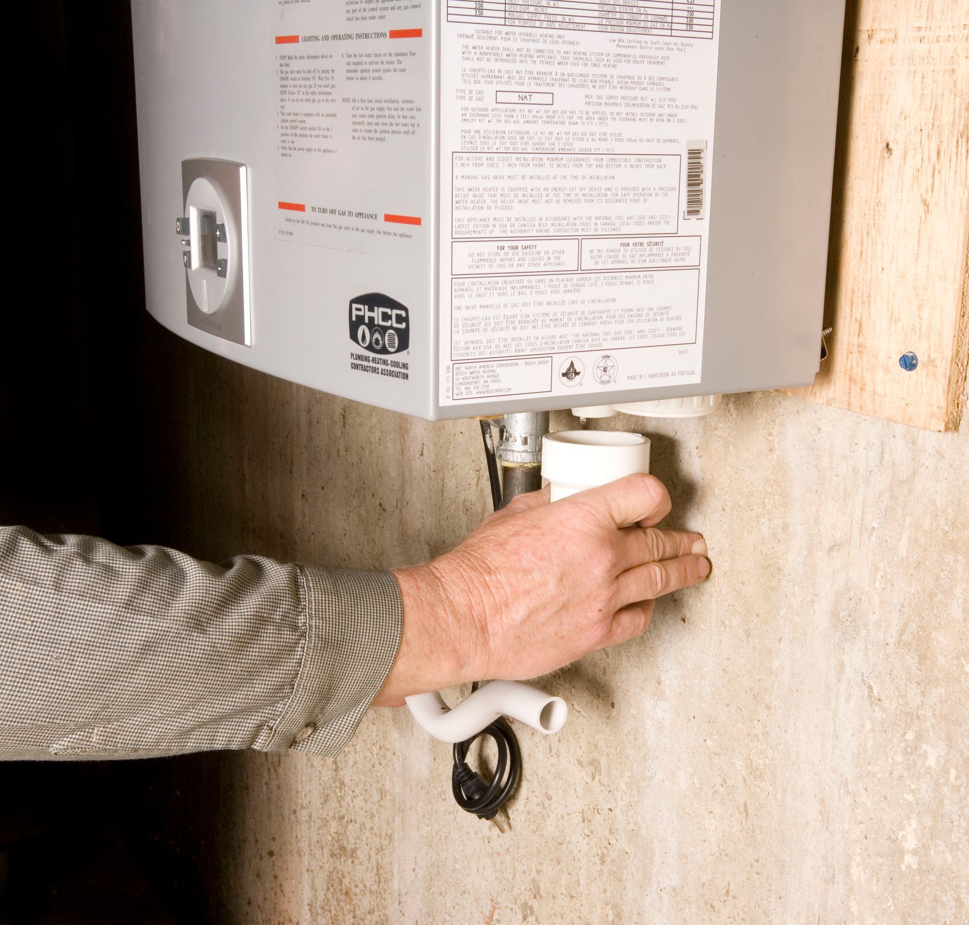 Person's hand near white water heater component; indoors. The water heater is mounted on a wall.