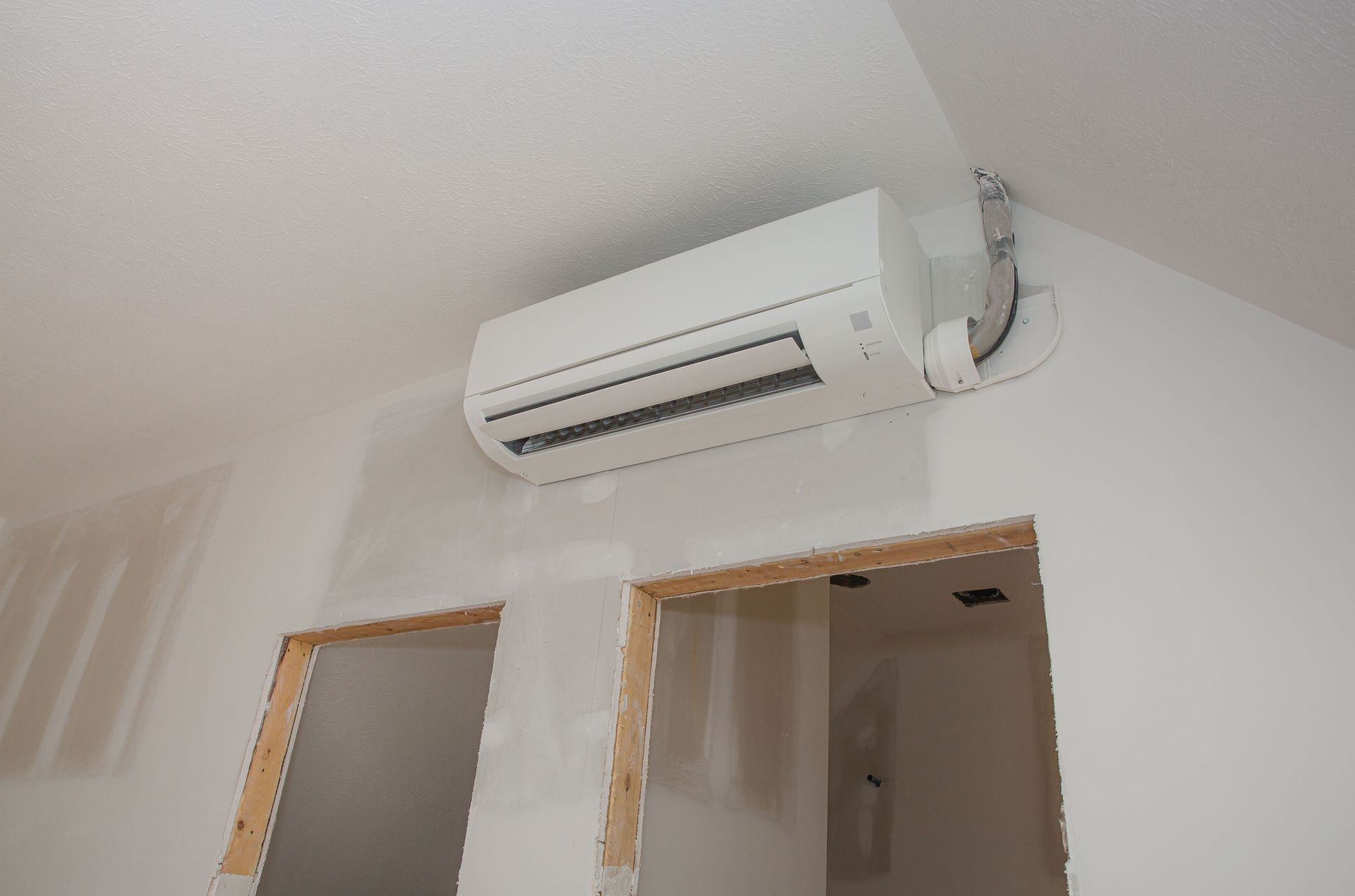 White air conditioning unit mounted on a white wall in a room under construction, with open doorways.