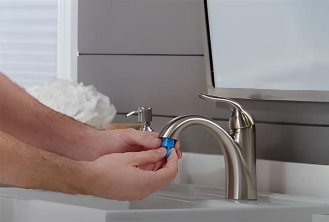 Hands attaching a blue aerator to a bathroom faucet, next to a soap dispenser and sink.