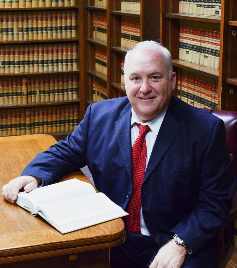 Man in suit, red tie, smiling, seated at desk with open book, in library setting.