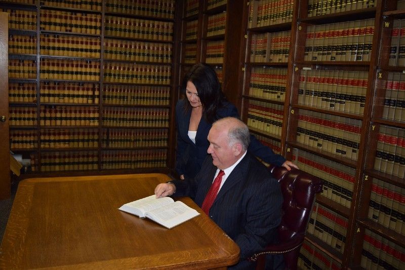 Lawyer in suit reviews a book at a desk as an associate looks on in a library with many law books.