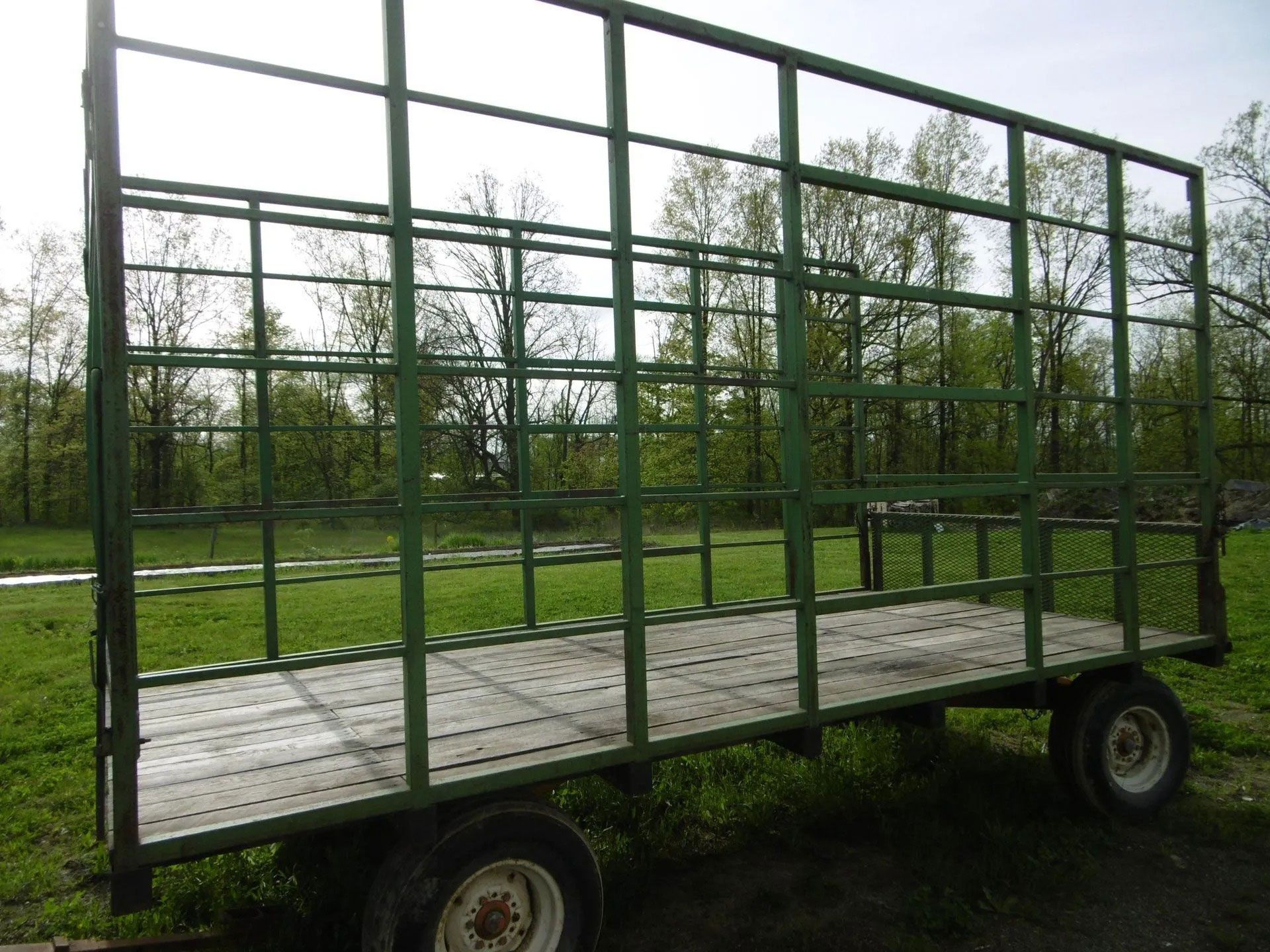 A green metal hay wagon frame with a wooden floor sits on a grassy field under a bright sky.
