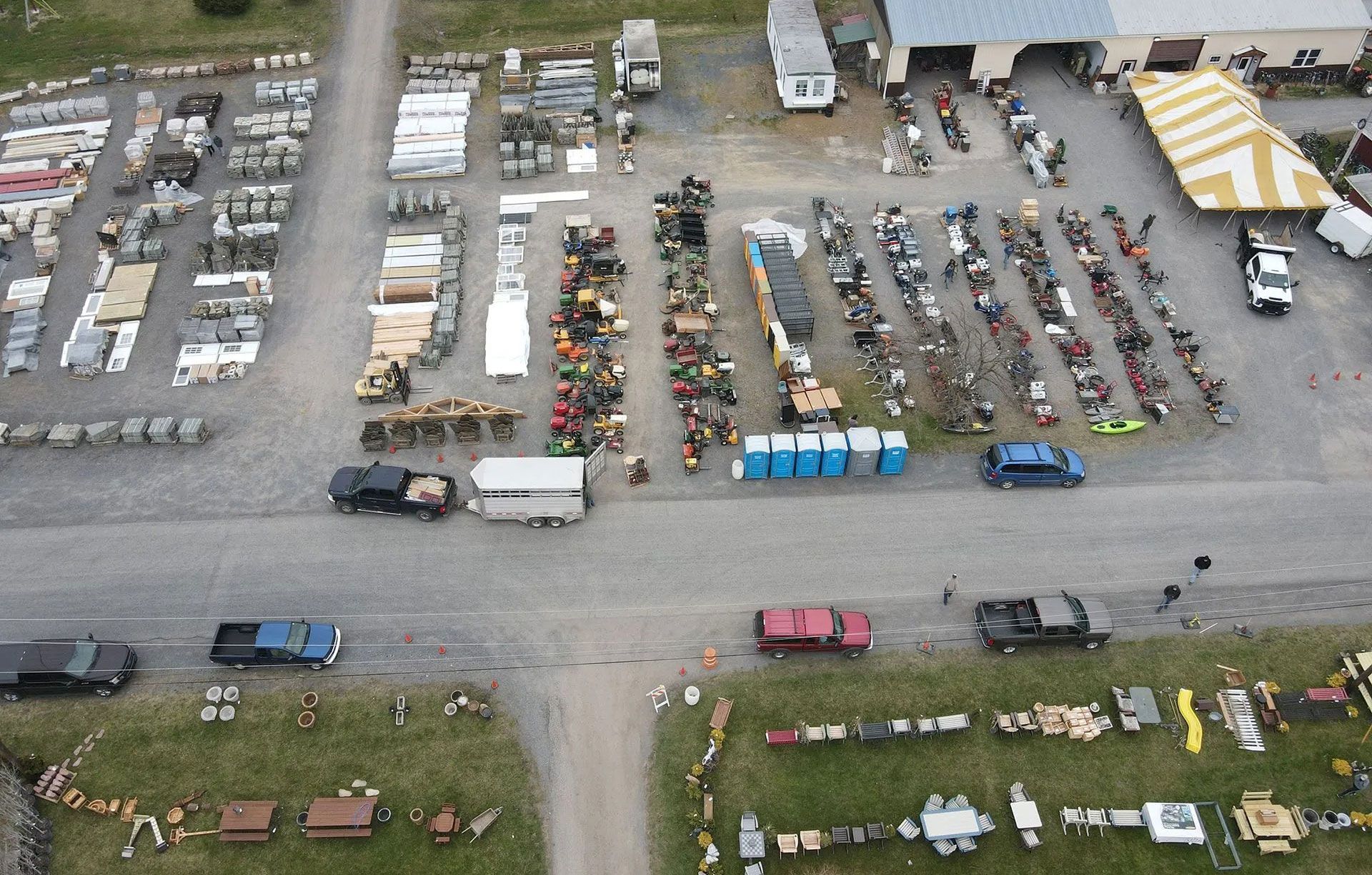 An aerial view of an outdoor auction or flea market with rows of items for sale, scattered vehicles, and portable toilets.