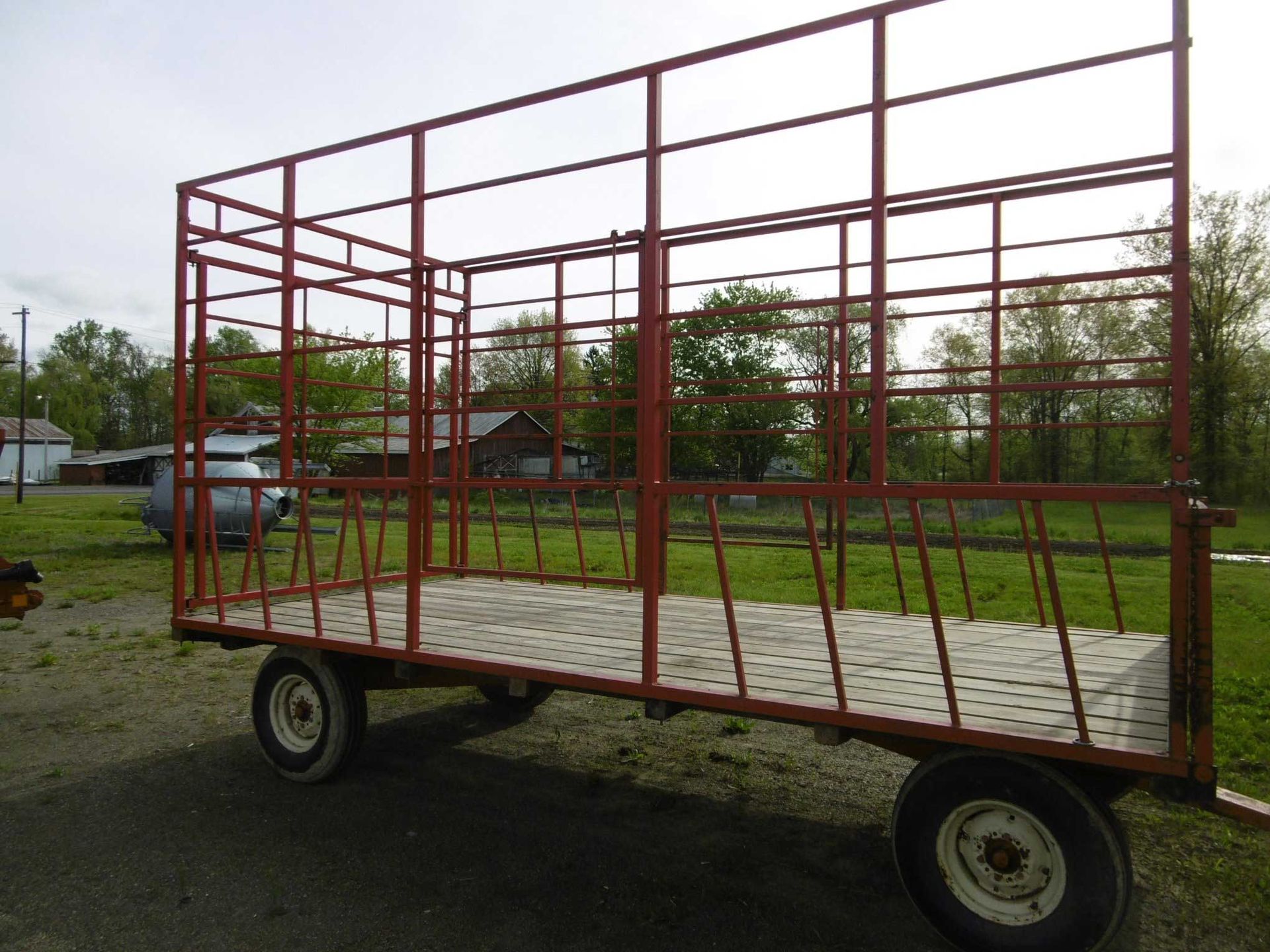 A red metal wagon frame with a mesh floor sits on a grass and gravel lot under a cloudy sky.