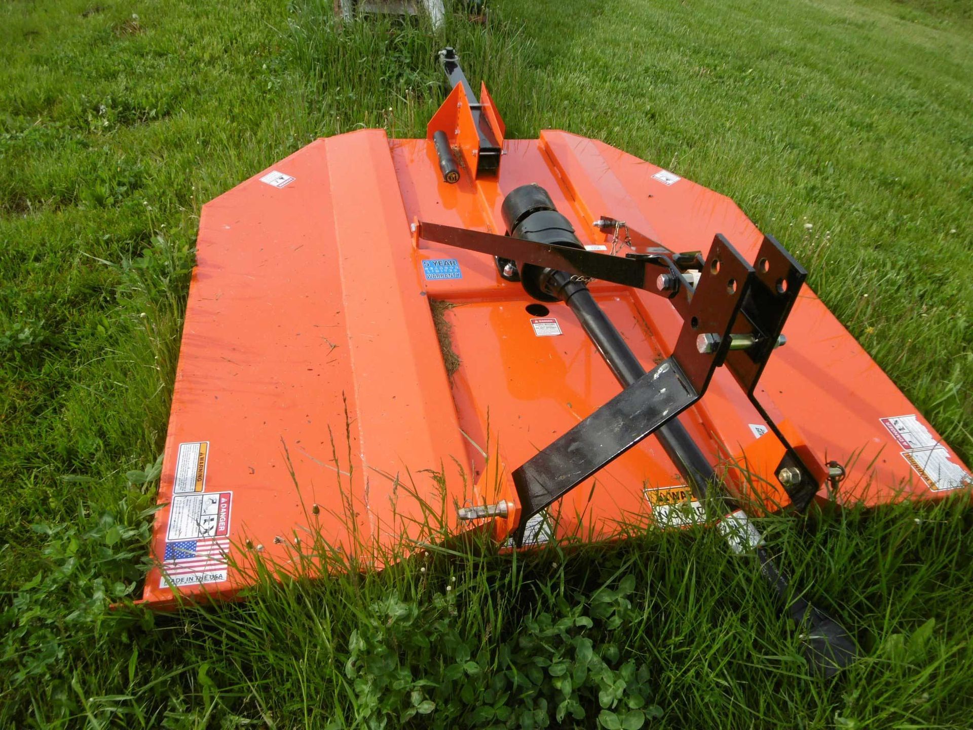 An orange rotary cutter, or brush hog, sitting in a grassy field, featuring a black metal 3-point hitch and drive shaft.